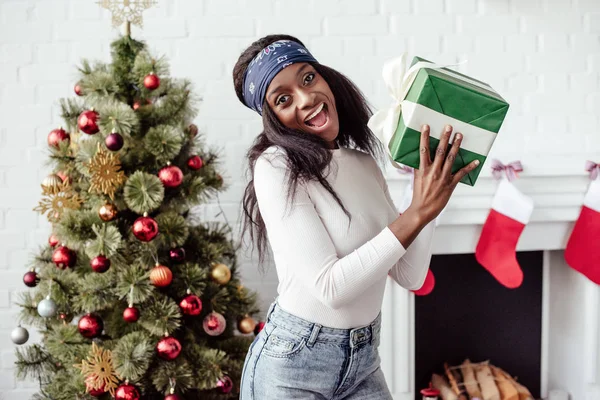 Aufgeregt attraktive afrikanisch-amerikanische Frau hält Weihnachtsgeschenk zu Hause und blickt in die Kamera — Stockfoto