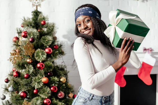 Happy beautiful african american woman holding christmas present at home and looking at camera — Stock Photo