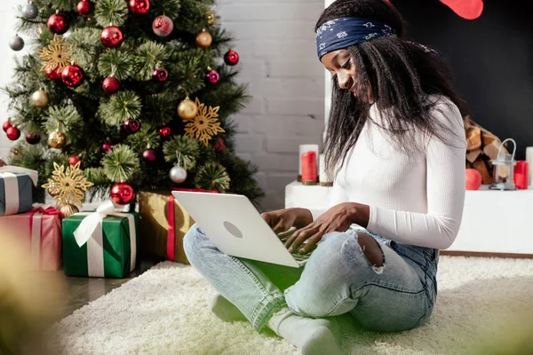 Attrayant afro-américaine femme en utilisant un ordinateur portable sur le sol à la maison, arbre de Noël dans le coin — Photo de stock