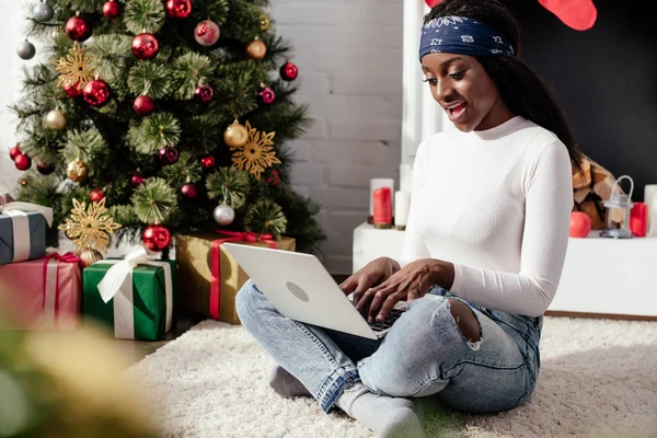Choqué attrayant afro-américain femme en utilisant un ordinateur portable sur le sol à la maison, concept de Noël — Photo de stock