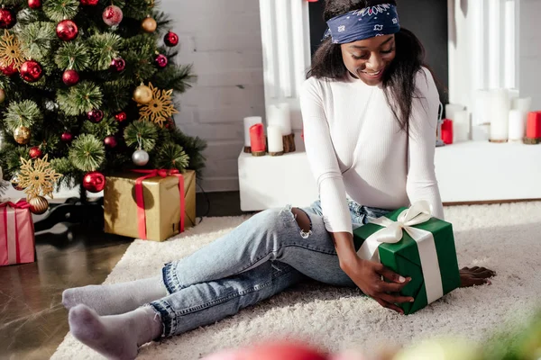 Heureuse femme afro-américaine attrayante regardant cadeau de Noël à la maison — Photo de stock