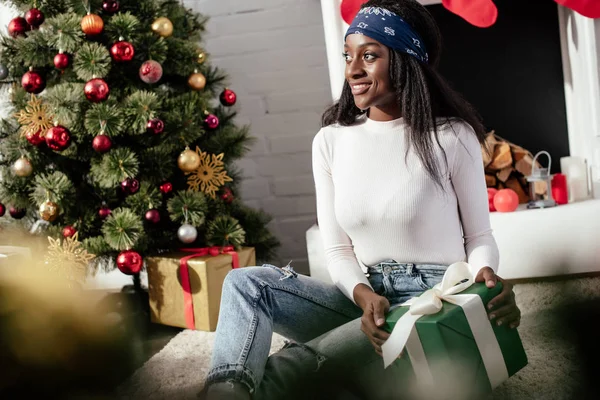 Atractiva mujer afroamericana sosteniendo la Navidad presente en casa y mirando hacia otro lado - foto de stock