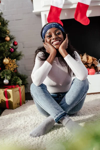 Sorridente attraente donna africana americana che poggia il mento sulle mani e guarda la fotocamera a casa, concetto di Natale — Foto stock