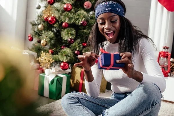 Emocionado atractiva mujer afroamericana apertura de la caja de regalo de Navidad en casa - foto de stock