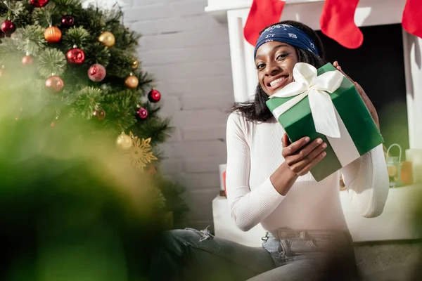 Aufgeregt attraktive afrikanisch-amerikanische Frau hält Weihnachtsgeschenk zu Hause und blickt in die Kamera — Stockfoto