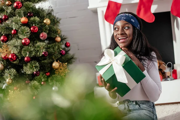 Excited attractive african american woman holding christmas present at home and looking away — Stock Photo