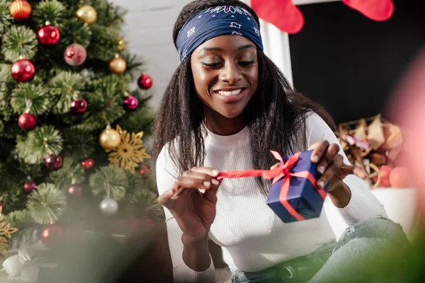 Selektiver Fokus der lächelnden attraktiven afrikanisch-amerikanischen Frau, die zu Hause das Weihnachtsgeschenk öffnet — Stockfoto