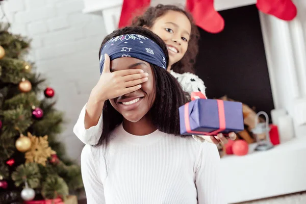 Sorrindo afro-americana filha presente natal presente para a mãe e fechar os olhos para surpresa em casa — Fotografia de Stock