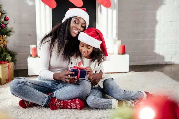 Sonriente afroamericana madre en santa hat presentando regalo a hija en casa y mirando cámara - foto de stock
