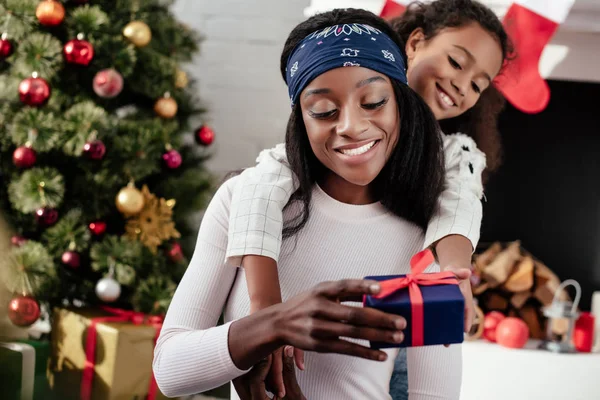 Hija afroamericana abrazando y regalando navidad presente a la madre en casa - foto de stock