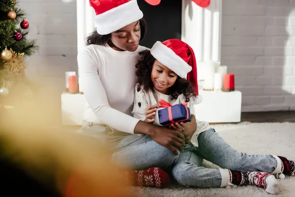 Selective focus of african american mother in santa hat presenting gift to daughter at home — Stock Photo