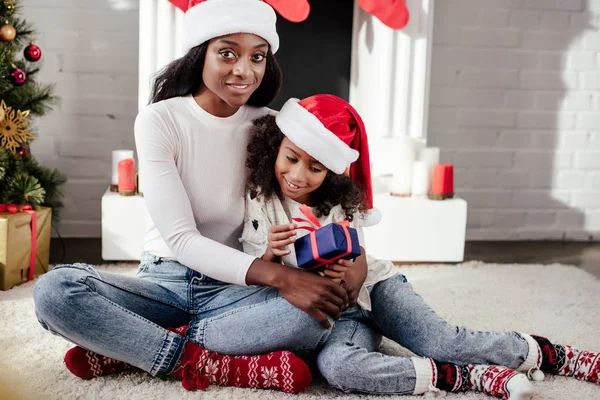 Afro-americana mãe em santa chapéu abraçando filha com presente em casa — Fotografia de Stock