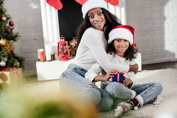 Улыбающаяся африканская американка обнимает маленькую дочь рождественским подарком в украшенной комнате дома — стоковое фото