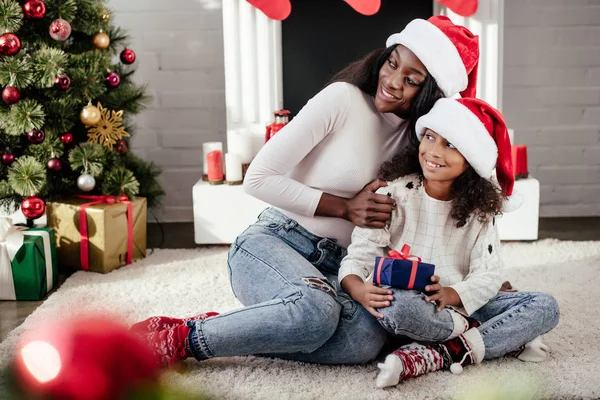 Sorrindo mulher afro-americana abraçando pequena filha com presente de Natal em quarto decorado em casa — Fotografia de Stock