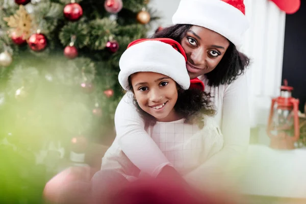 Афроамериканська мати в капелюсі Санта Клауса обіймає маленьку доньку в декорованій кімнаті вдома — стокове фото