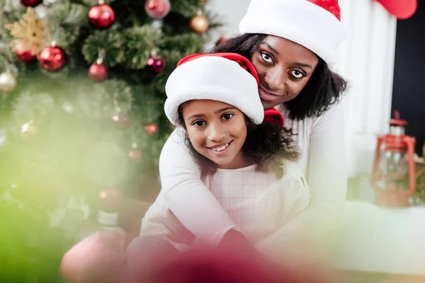 Afrikanisch-amerikanische Mutter mit Weihnachtsmann-Hut umarmt kleine Tochter in dekoriertem Zimmer zu Hause — Stockfoto