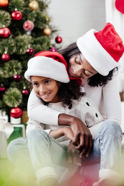 Africano americano madre en santa claus sombrero abrazando pequeña hija en decorado habitación en casa - foto de stock