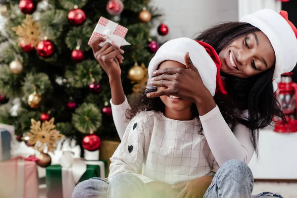 Souriant afro-américaine mère dans santa claus chapeau fille surprenante avec Noël présent à la maison — Photo de stock