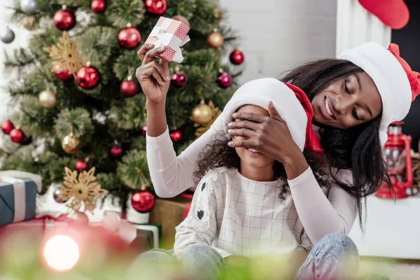 Souriant afro-américaine mère dans santa claus chapeau fille surprenante avec Noël présent à la maison — Photo de stock