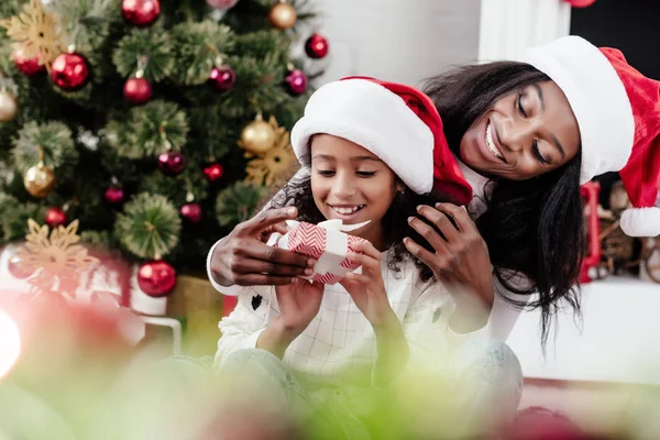 Happy african american woman and daughter with gift in santa claus hats at home, newy year and christmas concept — Stock Photo