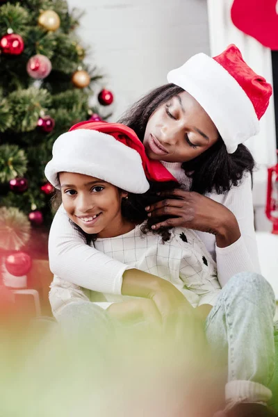 African american mother in santa claus hat hugging little daughter in decorated room at home — Stock Photo