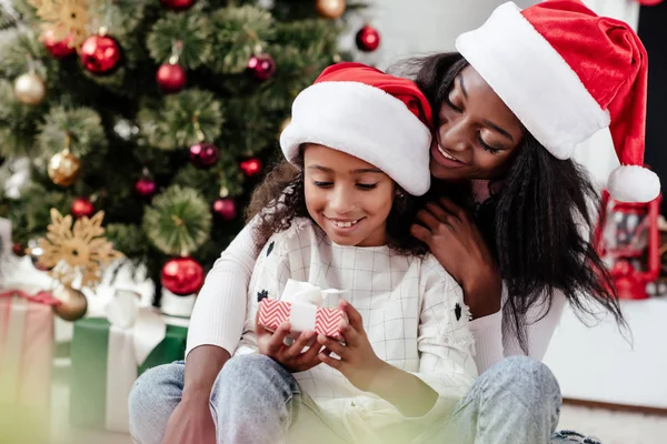 Happy african american woman and daughter with gift in santa claus hats at home, newy year and christmas concept — Stock Photo