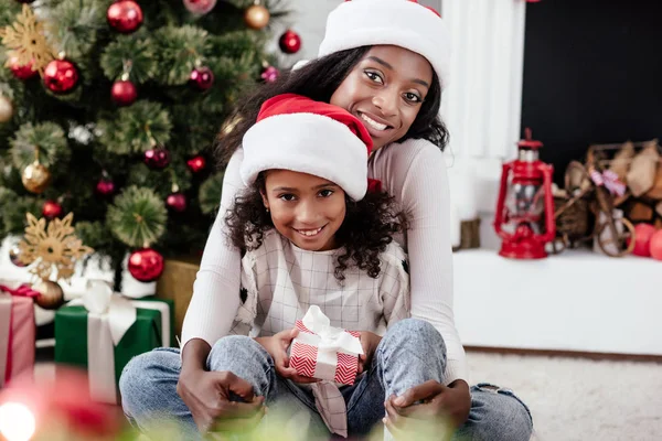 Happy african american woman and daughter with gift in santa claus hats at home, newy year and christmas concept — Stock Photo