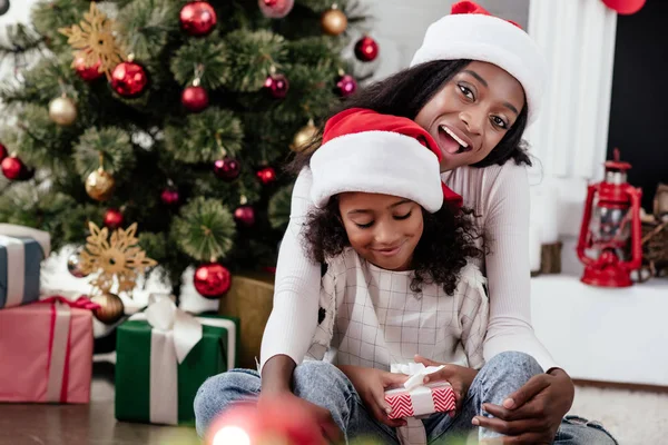 Happy african american woman and daughter with gift in santa claus hats at home, newy year and christmas concept — Stock Photo