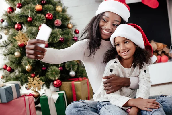 Sorridente afro-americano mãe e filha em santa claus chapéus tomando selfie no smartphone em casa — Fotografia de Stock