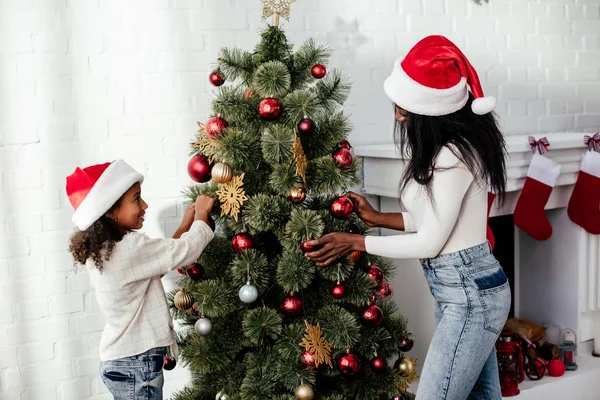 African american mother and daughter in santa claus hats decorating christmass tree together at home — Stock Photo