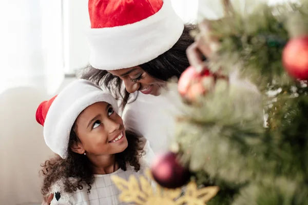 Glückliche afrikanisch-amerikanische Familie in Weihnachtsmannhüten, die zu Hause gemeinsam den Weihnachtsbaum schmücken — Stockfoto