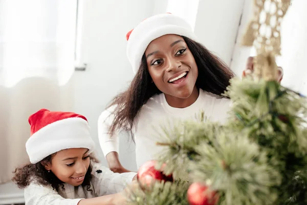Retrato da feliz família afro-americana em chapéus de santa claus decorando árvore de natal juntos em casa — Fotografia de Stock