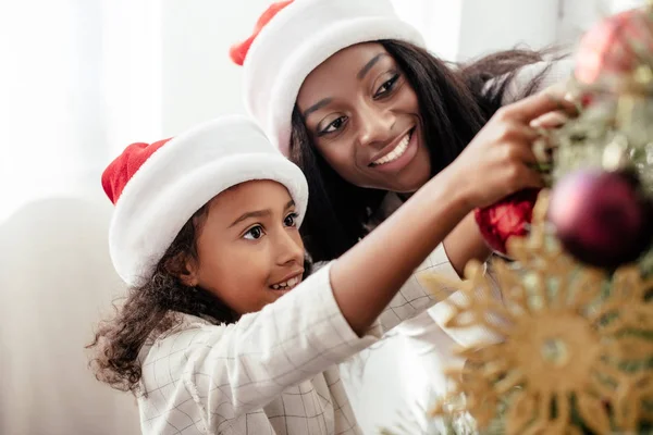 Africano americano madre e hija en santa claus sombreros decoración christmass árbol juntos en casa - foto de stock