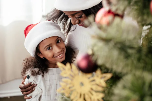 Glückliche afrikanisch-amerikanische Familie in Weihnachtsmannhüten, die zu Hause gemeinsam den Weihnachtsbaum schmücken — Stockfoto