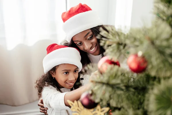 Portrait of happy african american family in santa claus hats decorating christmas tree together at home — Stock Photo