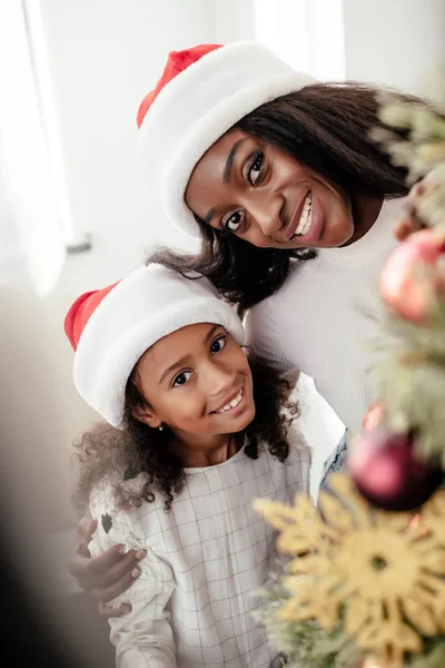 Retrato de la feliz familia afroamericana en sombreros de Santa Claus decorando el árbol de Navidad juntos en casa - foto de stock