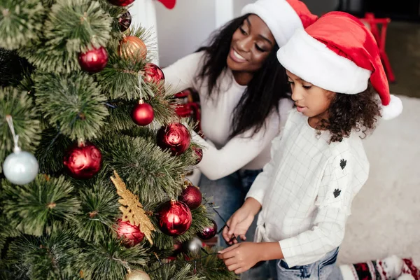 African american mother and daughter in santa claus hats decorating christmass tree together at home — Stock Photo