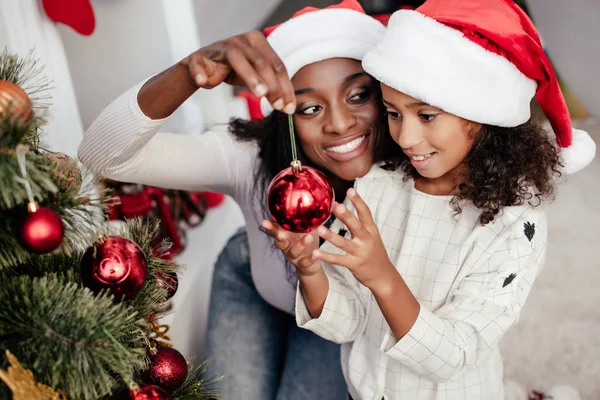 Sonriente afroamericana mujer en santa claus sombrero ayudar hija a decorar árbol de Navidad en casa - foto de stock