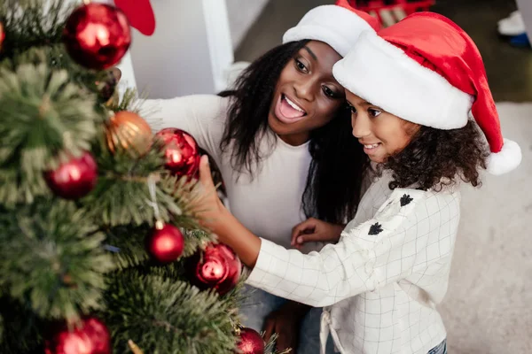 Sonriente afroamericana mujer en santa claus sombrero ayudar hija a decorar árbol de Navidad en casa - foto de stock