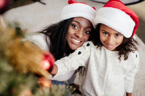 Smiling african american woman in santa claus hat helping daughter to decorate christmas tree at home — Stock Photo