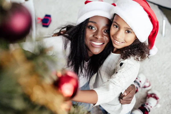 Lächelnde Afroamerikanerin mit Weihnachtsmann-Hut hilft Tochter, Weihnachtsbaum zu Hause zu schmücken — Stockfoto