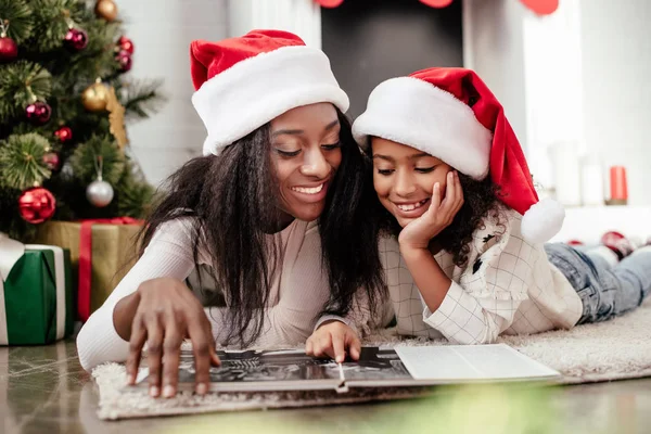Sorrindo família afro-americana em chapéus de santa claus olhando para álbum de fotos em quarto decorado para o Natal em casa — Fotografia de Stock