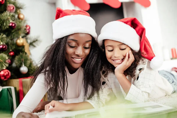 Lächelnde afrikanisch-amerikanische Familie mit Weihnachtsmann-Hüten beim Betrachten eines Fotoalbums im weihnachtlich dekorierten Raum zu Hause — Stockfoto