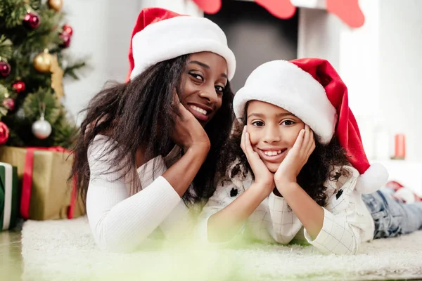 Alegre afroamericano familia en santa claus sombreros acostado en el suelo en habitación decorada para la Navidad en casa - foto de stock