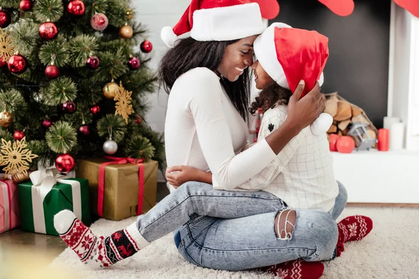 Glückliche afrikanisch-amerikanische Frau und Tochter in Weihnachtsmannhüten umarmen sich in dekorierten Raum für Weihnachten zu Hause — Stockfoto