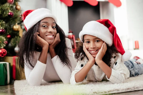 Веселая африканская американская семья в шляпах Санта-Клауса, лежащих на полу в украшенном помещении на Рождество дома — стоковое фото
