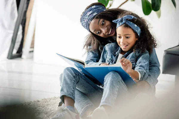 African american mother and smiling daughter looking at family photo album at home — Stock Photo