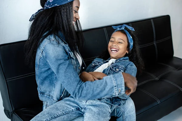 Happy african american woman having fun with daughter in similar clothing on sofa at home — Stock Photo