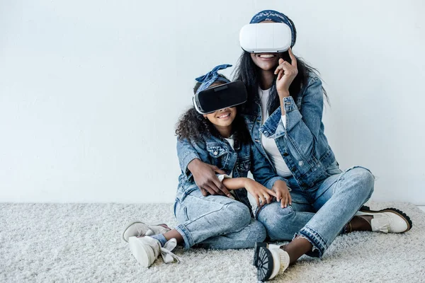 African american mother hugging daughter in similar clothing and vr headsets at home — Stock Photo