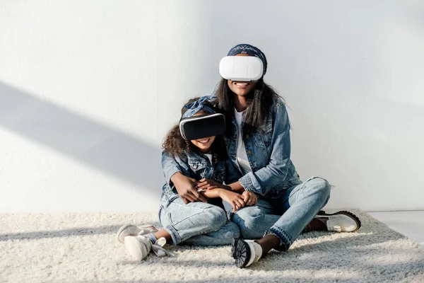 Afro-americana mãe abraçando filha em roupas semelhantes e fones de ouvido vr em casa — Fotografia de Stock